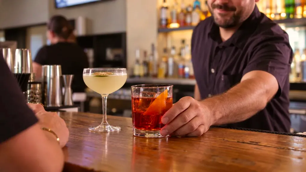 A bartender setting a cocktail on the bar next to a coupe glass